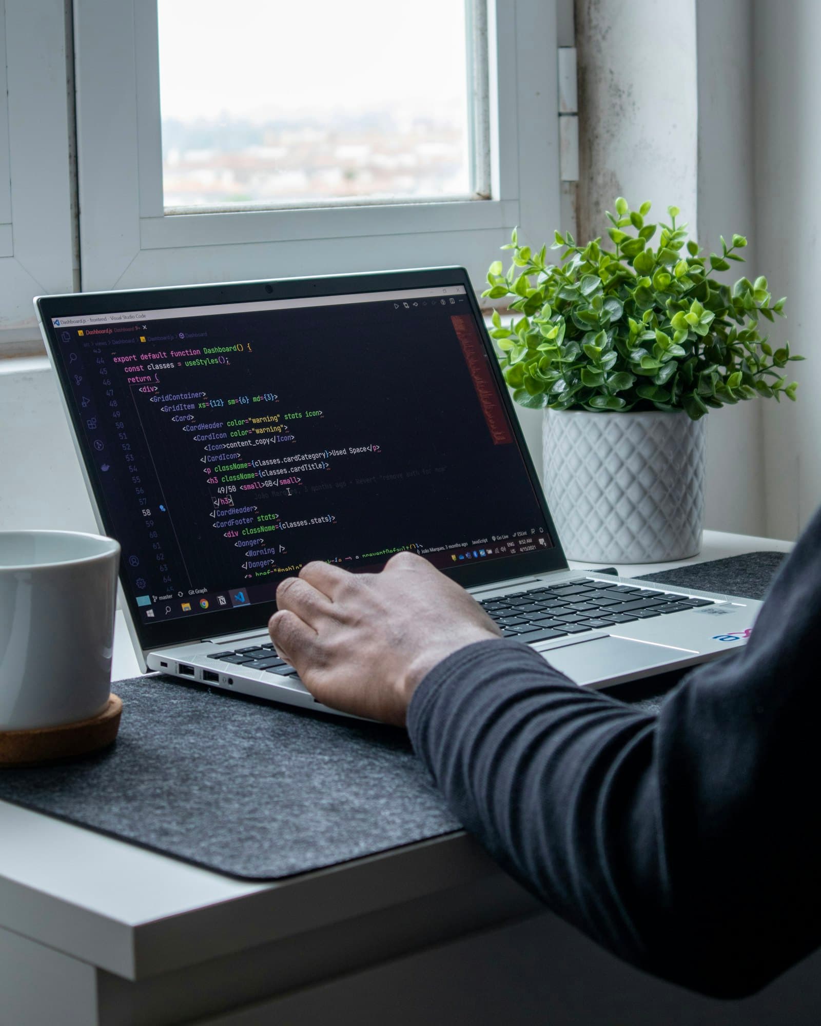 A developer typing code on a laptop by a window, a plant and a mug beside it