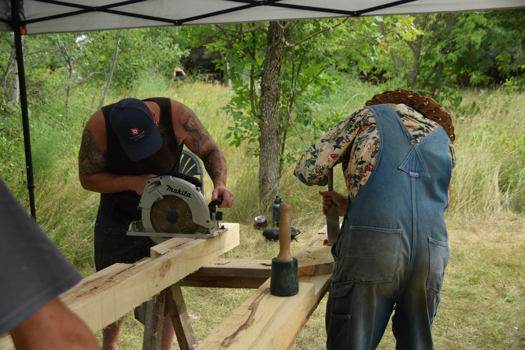Two people cutting and chiselling a timber beam outdoors under a canopy